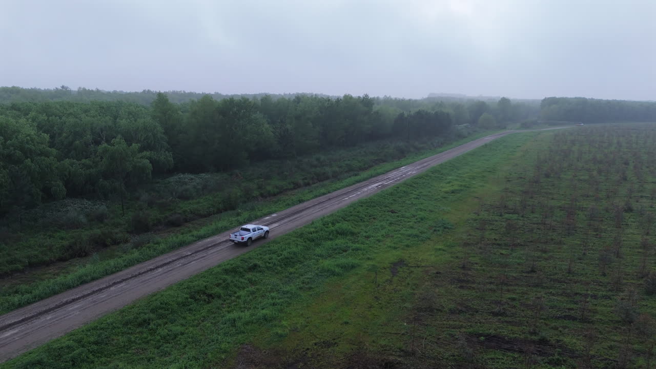 Aerial view of a white car driving on a dirt road through the countryside, surrounded by forest and fields on a cloudy day. Scenic travel and rural adventure.