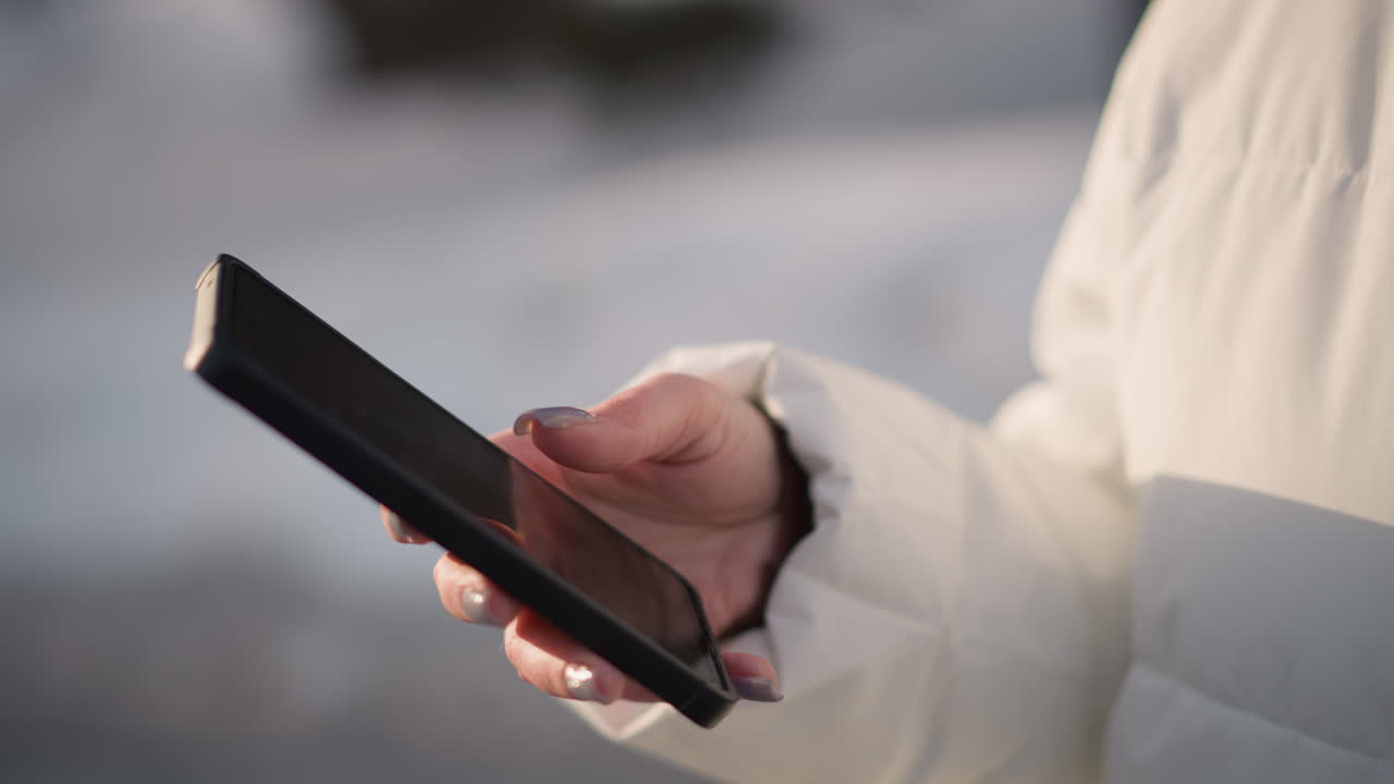 Student scrolling smartphone screen with manicured nails against white winter jacket in park while body sways, conveying digital lifestyle exploration under soft natural light breeze
