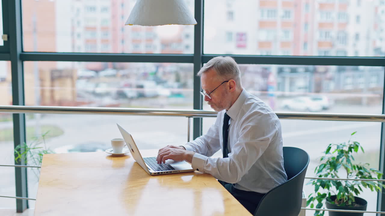 Side view of a senior man typing on a laptop. Successful businessman in white shirt and tie sitting at the desk and working near the window.