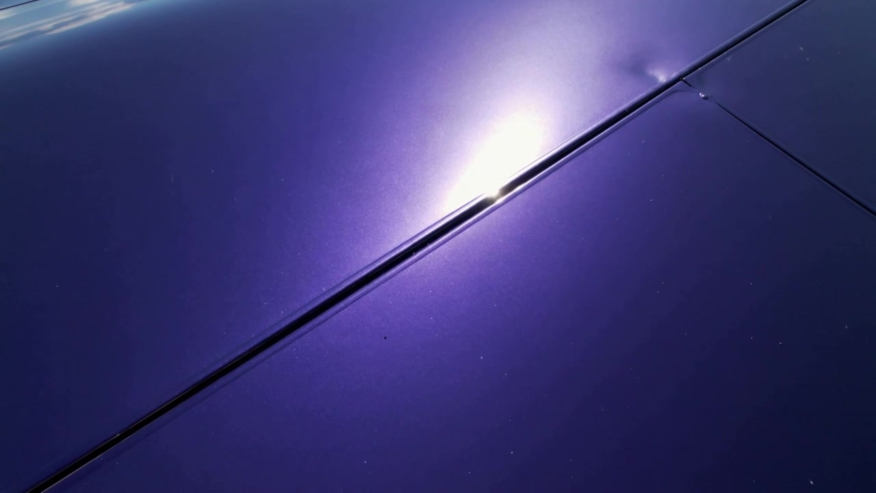 Câmera em movimento inspecionando o painel roxo de um carro na entrada da garagem, revelando um amassado acima da emenda com o brilho do sol.