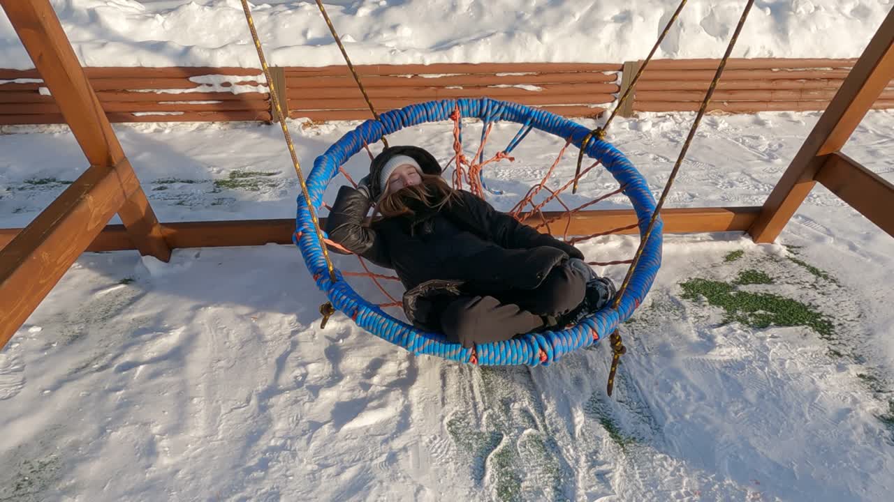 una adolescente cansada con ropa exterior negra disfruta balanceándose y relajándose acostada en una cuerda de mimbre en un swing redondo en un día de invierno. nieve en el fondo.