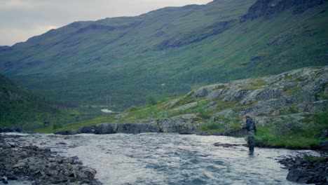 Man-fly-fishing-in-stream-by-green-mountains-in-Sweden,-wide-view