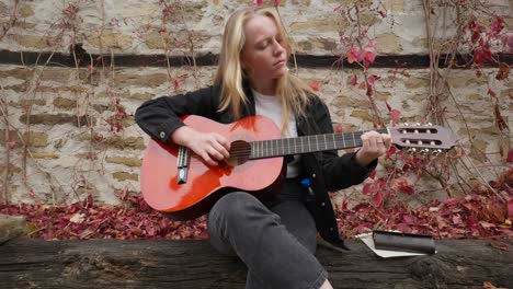 Beautiful-performer-plays-guitar,-autumn-foliage-stone-wall-scene-push-shot