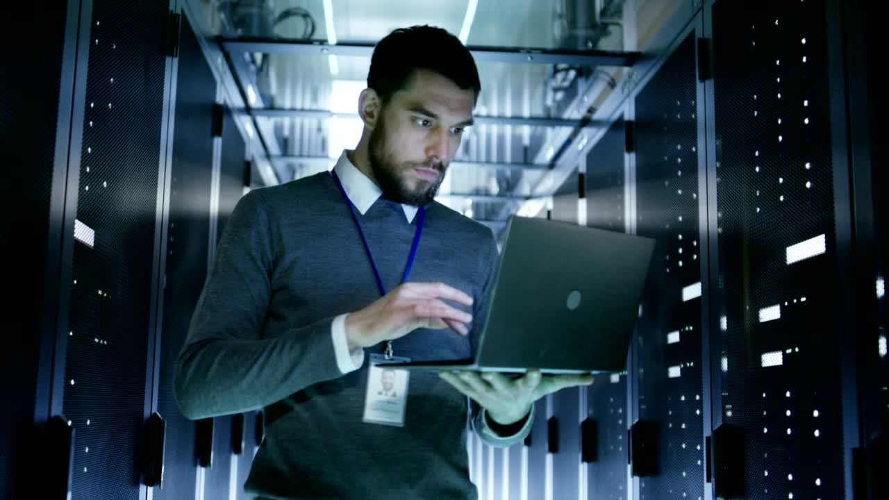 IT Technician Works on Laptop in Big Data Center full of Rack Servers. He Runs Diagnostics and Maintenance.