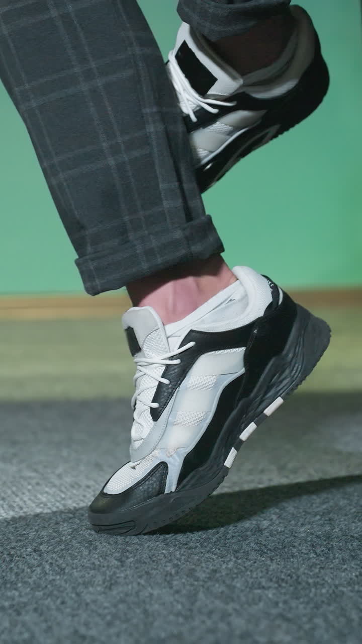 회색 카 바지를 입은 세련된 색과 검은색 스니커를 입은 남성 다리의 클로즈업. 회색 카페트 바닥에서 발을 약간 들어 올려 현대적인 패션 성명을 만니다.