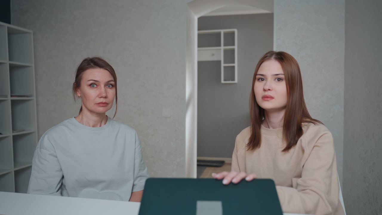 Two caucasian women at desk, tutor correcting students grammar using laptop, soft lit home office with shelves and plant, notebook and pen on table, gesturing explanation, attentive expressions