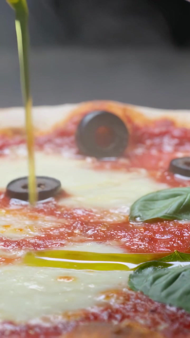 Video verticale: Versare olio da una bottiglia su una pizza napoletana con olive e basilico su un bancone lucido.