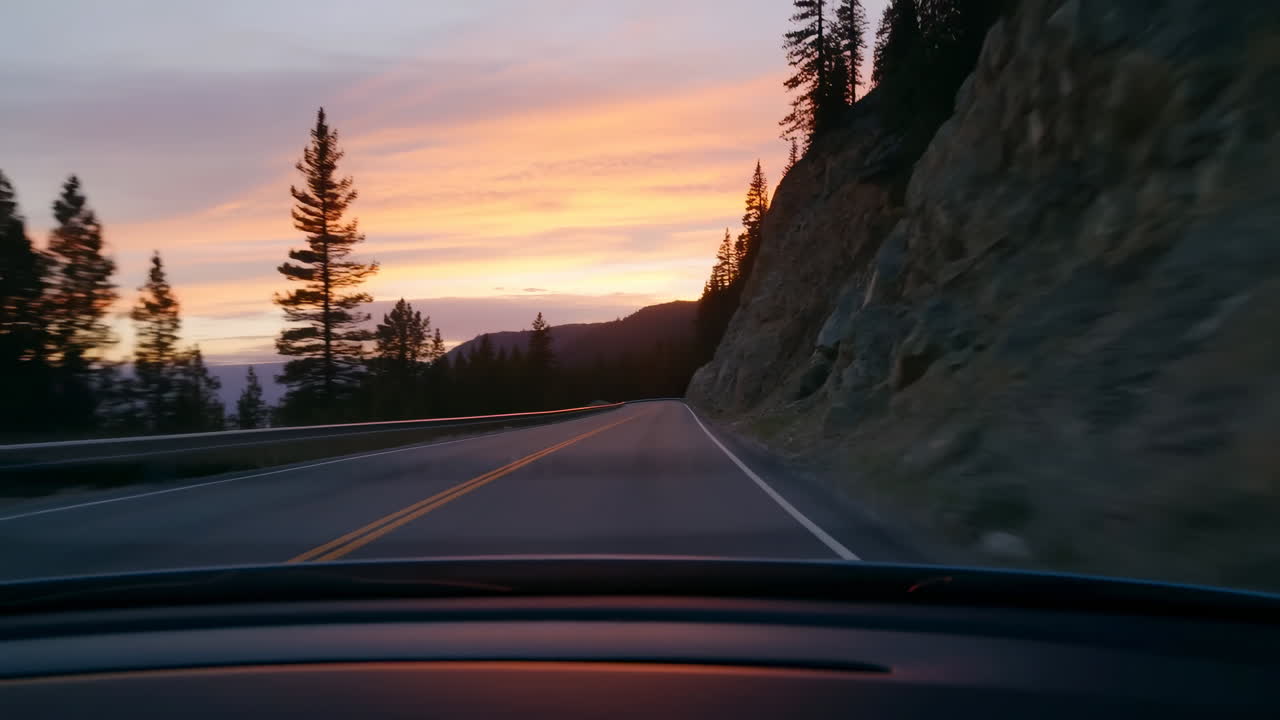 Bjergvej ved solnedgang fra bilens instrumentbræt, snoet kurve krammer klippefyldt skrænt, høje fyrretræer silhuetteret