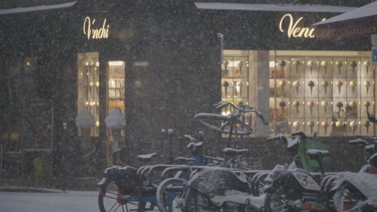 Biciclette parcheggiate fuori da una vetrina parigina illuminata calorosamente sono coperte di neve fresca mentre i fiocchi continuano a cadere al crepuscolo in inverno