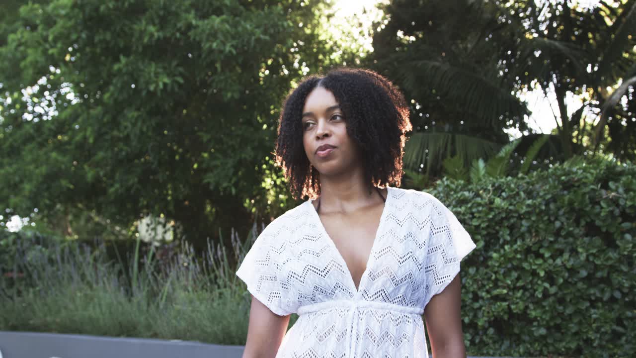 Mujer afroamericana moviendo las manos nerviosamente, bajando los brazos, mirando a la derecha en un jardín con un vestido blanco.