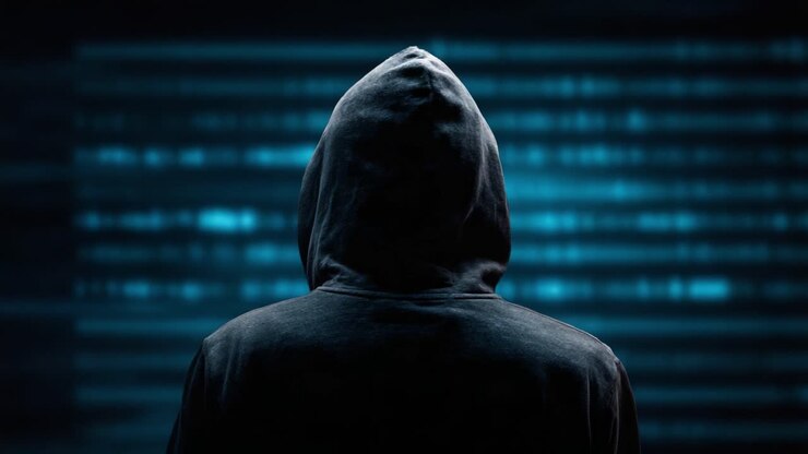 Una figura incappucciata in una felpa scura si staglia contro uno sfondo di codice digitale, evocando mistero e un'atmosfera di cyber-intrighi e violazioni della sicurezza in agguato nell'ombra.