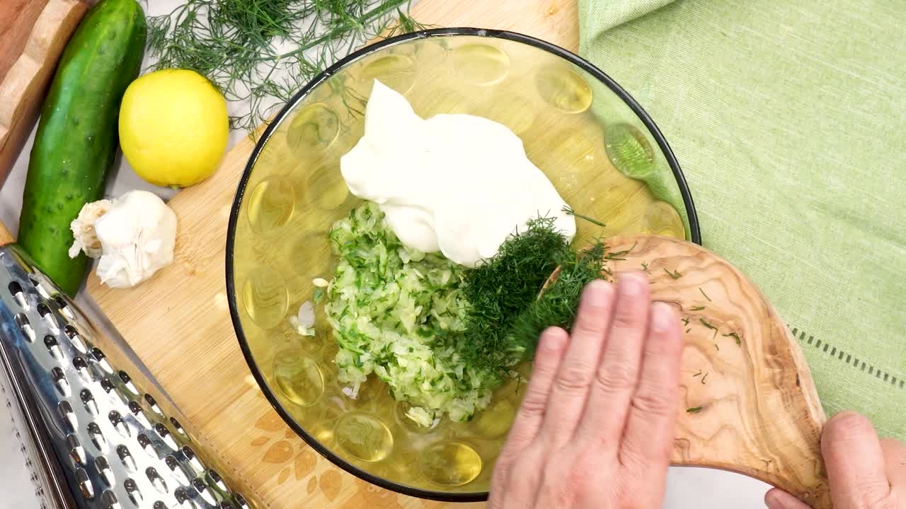 Making Tzatziki Greek or Mediterranean sauce or dip for a party. Dip or sauce in a green bowl on a wooden cutting board with dill, green napkin and a grater. Adding dill to the Tzatziki sauce.