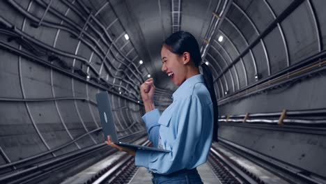 side view of asian female with her laptop in underground subway tunnel, she raises her fist up with screaming goal after typed on the laptop