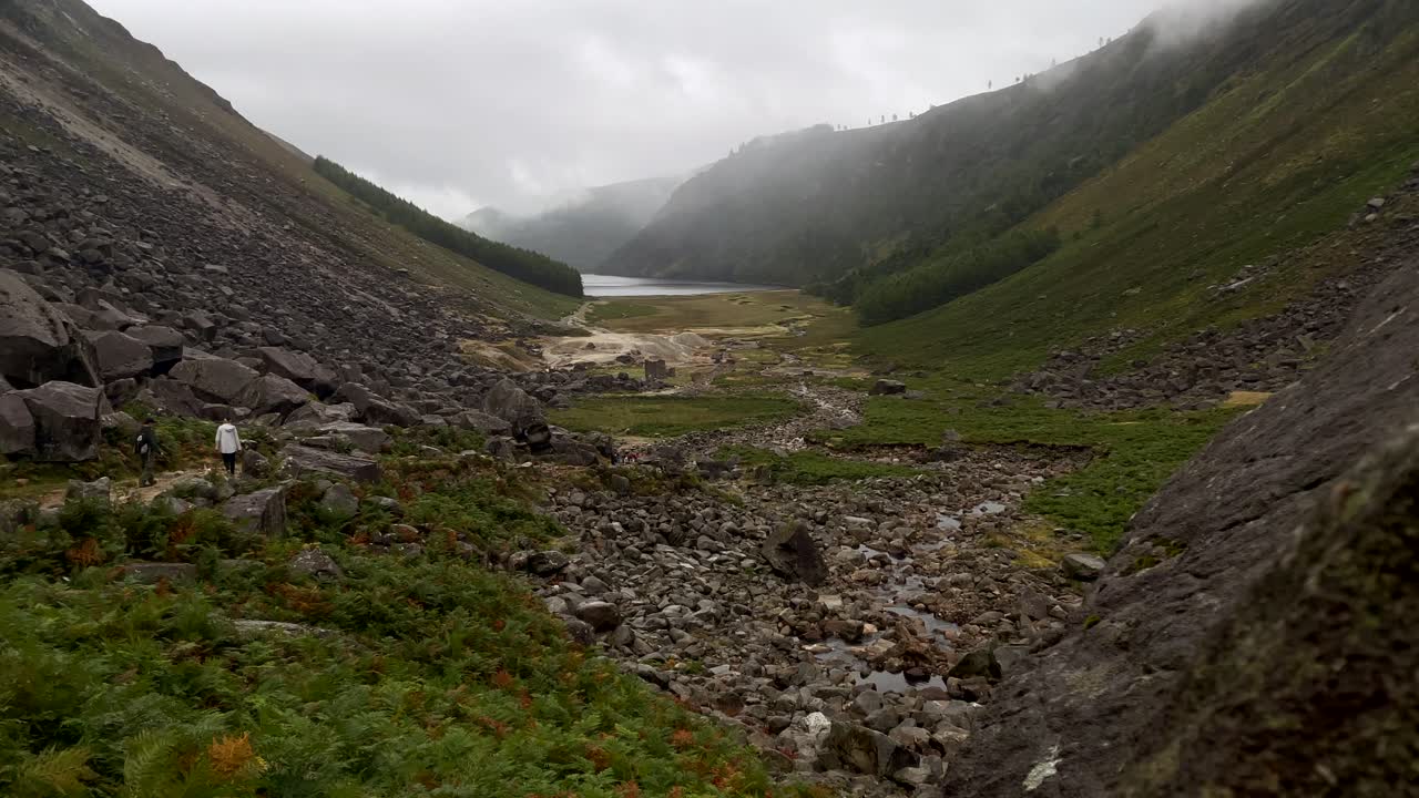 Bidikan yang menampakkan pendaki gunung di pegunungan berbatu Irlandia selama hari berawan dan hujan - Bidikan panning pemandangan perbukitan indah dengan danau alami di latar belakang