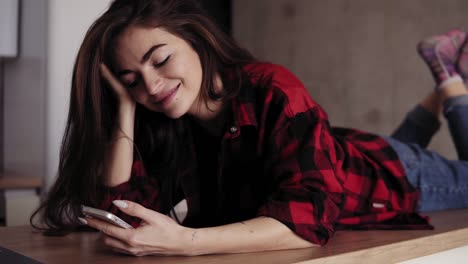 Attarctive-brunette-girl-is-dreaming-and-smiling-about-something-while-lying-on-kitchen-table-surface-and-texting-someone.-Close-up-footage.