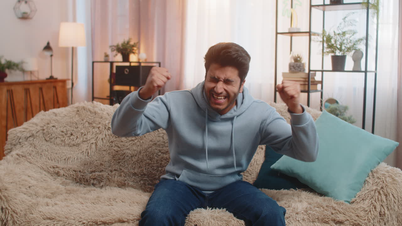 Happy indian young man celebrating success and victory shouting while sitting on sofa at home