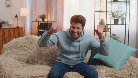 happy indian young man celebrating success and victory shouting while sitting on sofa at home