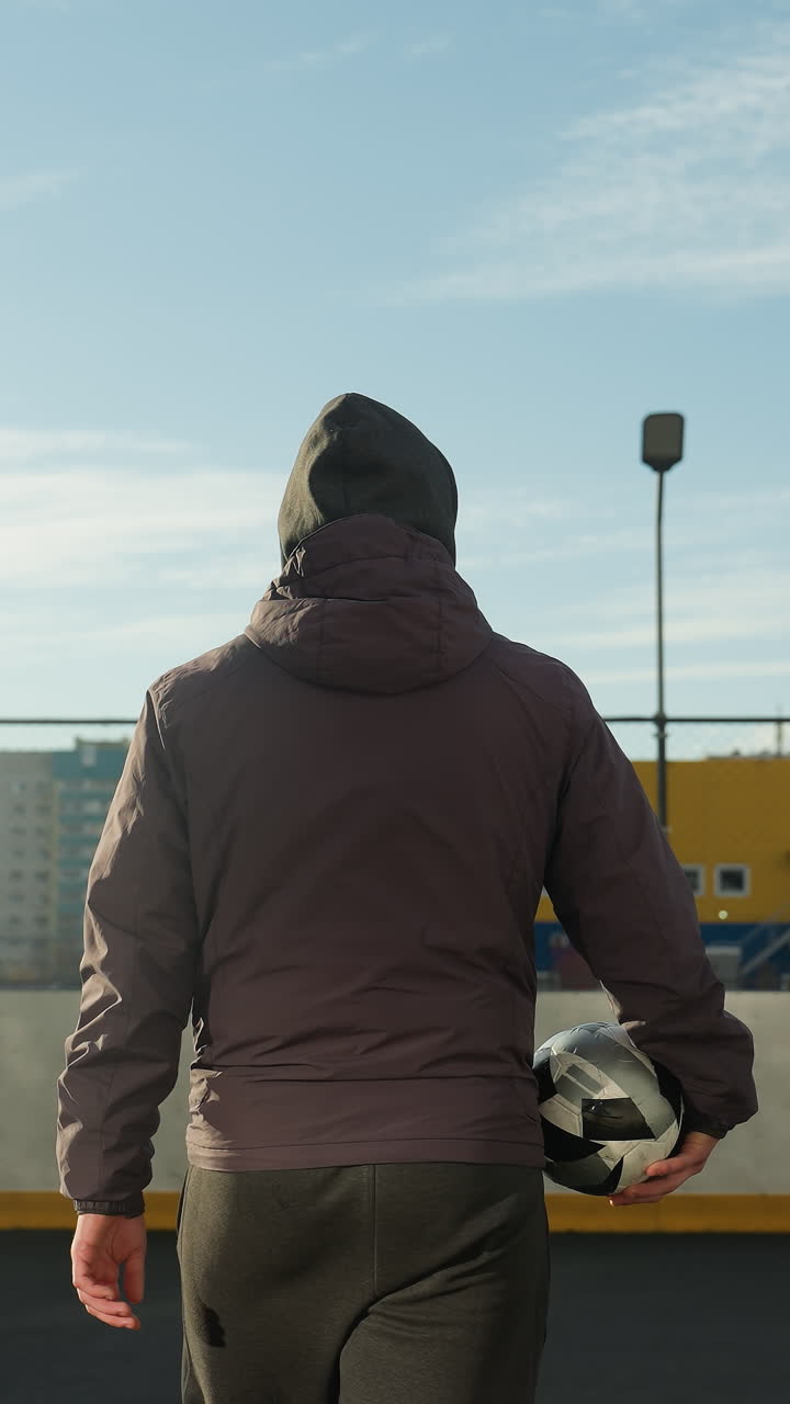 도시 스포츠 경기장에서 멀리 걸어가는 남자의 뒷면, 도시 풍경과 노란색 저장 컨테이너를 배경으로 게이트를 향해 축구 공을 들고