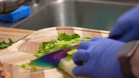 hands wearing rubber gloves with fresh lettuce cut into shreds at wooden board. - closeup shot
