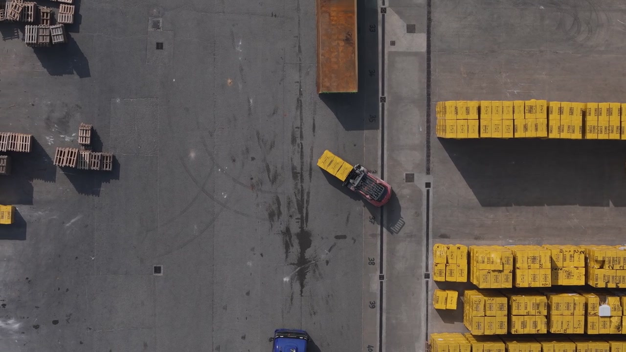 Forklift transporting concrete blocks toward truck trailer inside industrial storage yard in Po Valley Italy during top down drone tracking aerial shot focused on logistics activity