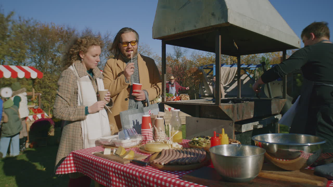 Couple Stand Near Grill at Farmers Market Couple at Autumn Fair or Farmers Markets Adult Man Cuts and Cooks Meat Steak on Grill Holder of Point of Sale system Outdoors Picnic People Walking on Background Slow Motion