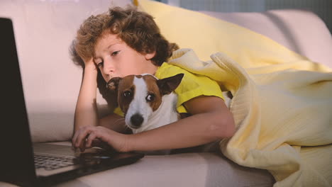 Blond Boy With Curly Hair Using Computer While Lying With His Dog On The Sofa