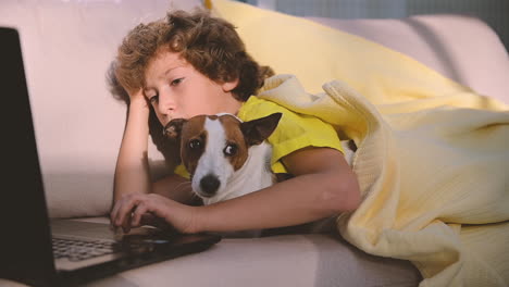 niño rubio con el pelo rizado usando la computadora mientras está acostado con su perro en el sofá