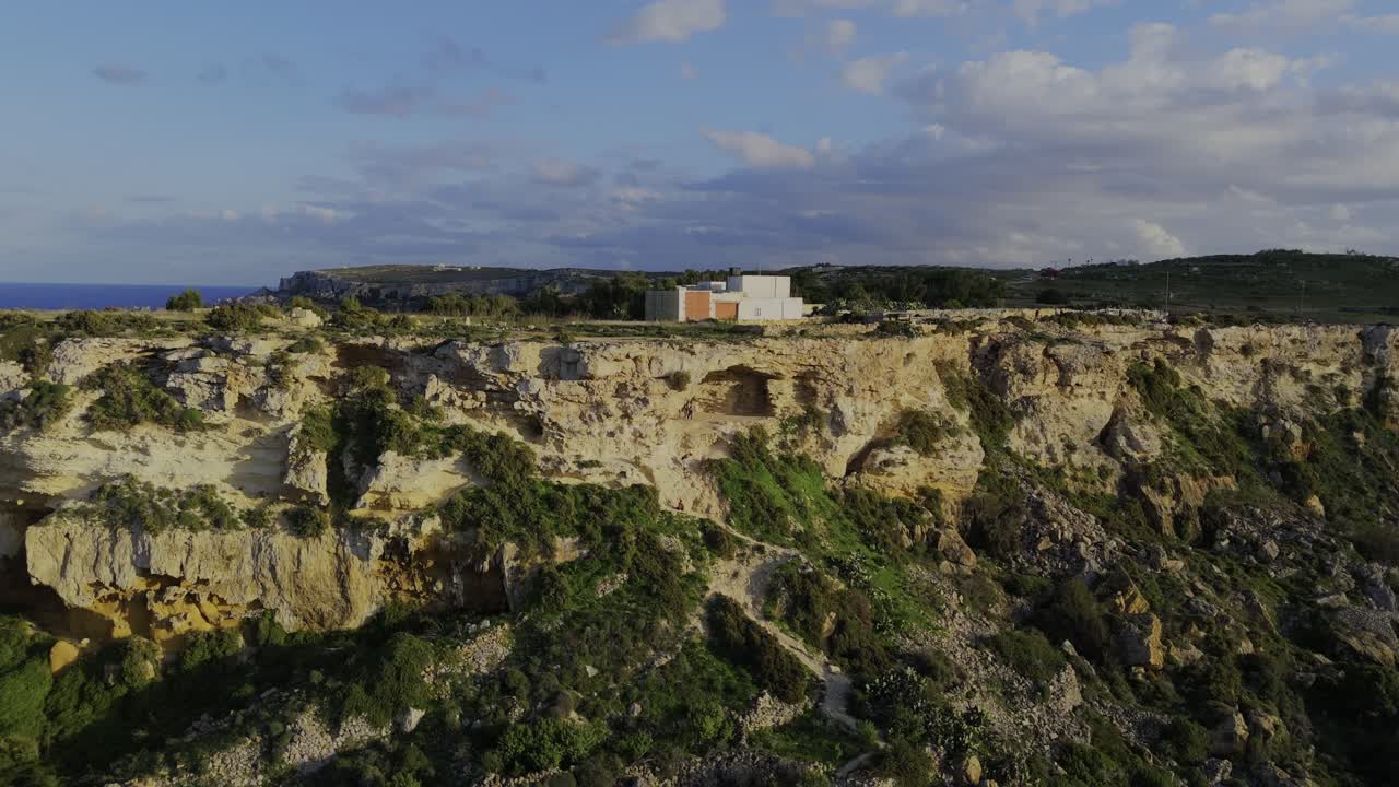 A smooth drone descent over Gozo reveals Mixta Cave glowing in warm sunset light, with rugged cliffs, blue sky, soft clouds and Mediterranean colors