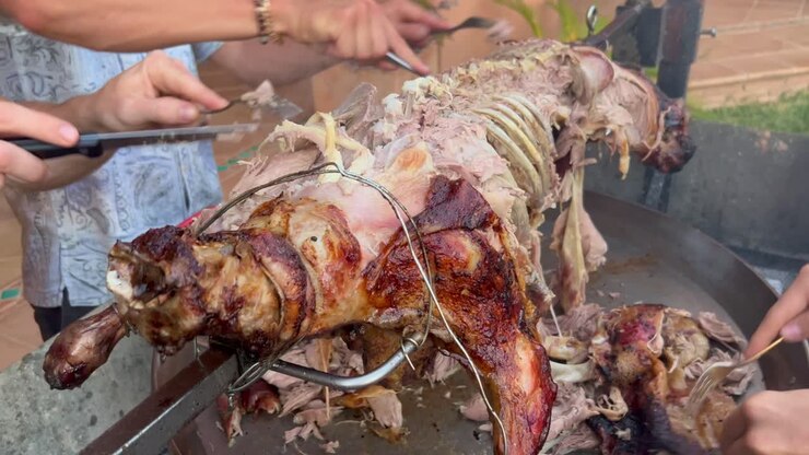 Tangan pria memotong seekor domba utuh dengan garpu seperti orang yang makan lahap di atas tusuk sate logam dengan kulit renyah keemasan, waktu memanggang barbekyu domba selama musim panas, rekaman 4K.