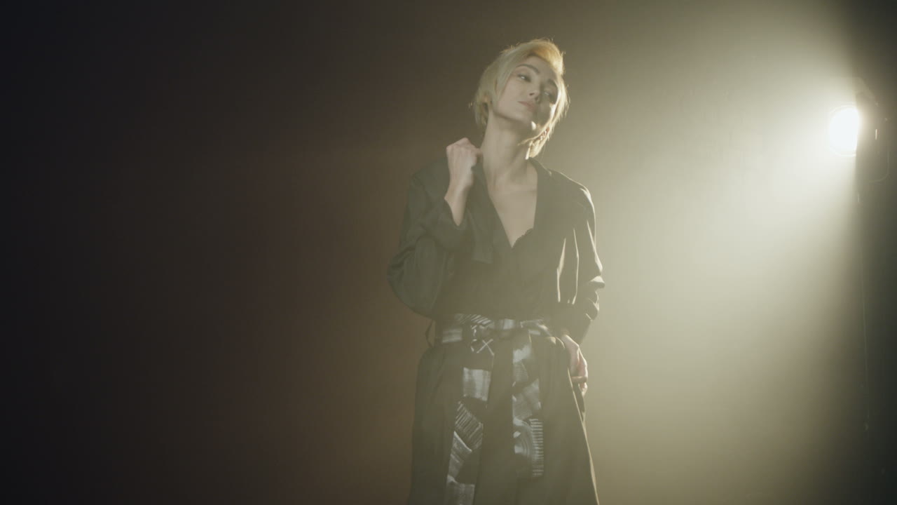 사진 작가가 그녀의 사진을 찍는 동안 스포트라이트와 함께 어두운 스튜디오에서 포즈를 취하는 백인 젊은 여성 모델