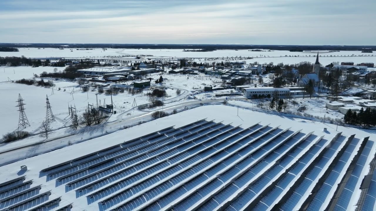 Dron letící pomalu dozadu nad panely solární elektrárny v řadě na zasněžených polích, v pozadí je obec Haljala, Estonsko