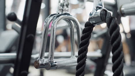 close up of chrome triangle grip and black rope attachment hanging on cable machine, with textured handle and blurred gym-goer in background, highlighting accessory details for strength training
