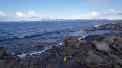 Toma-Aérea-Hacia-Atrás-De-Un-Turista-Sentado-Entre-Rocas-Para-Tomar-Fotografías-De-Gaviotas-Volando-Sobre-El-Océano-Cerca-De-La-Orilla