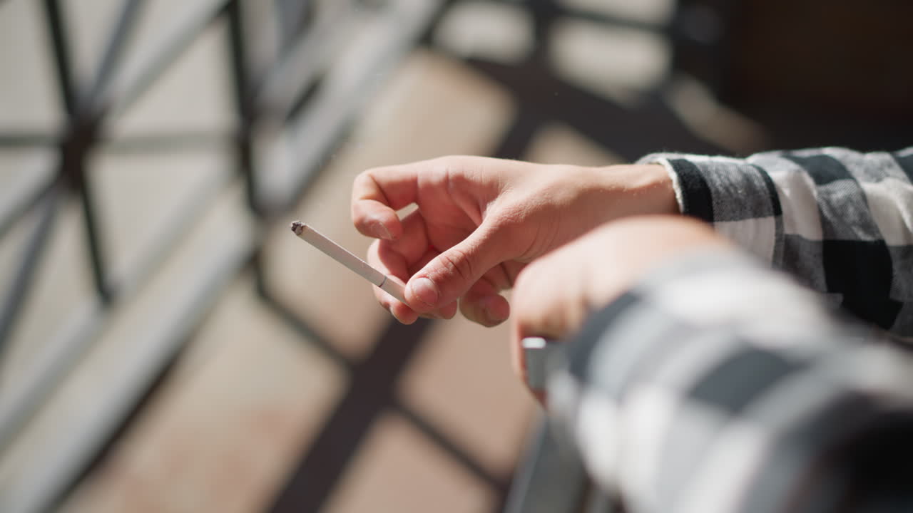 hand view of smoker tapping lit cigarette against iron railing twice to drop ash in warm sunlight with blurred urban background checkered shirt sleeve flickering in breeze