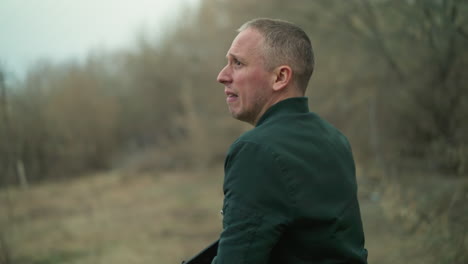 a close view of a man in a green jacket, with a tense expression, reaching into his pocket to draw a pistol while looking over his shoulder in a forested area