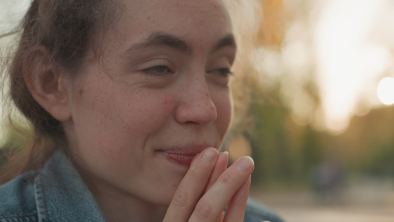 Thoughtful lips and clasped hands, young white woman imagining marriage and family in golden park light, closeup on freckled skin, sincere hopeful expression, quiet dreamer moment
