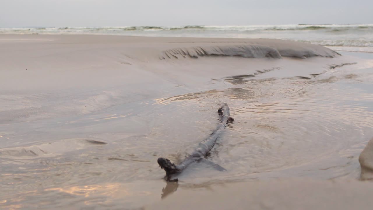 Low angle footage of water flowing and making its way to ocean water on a sandy beach during a cloudy day in slow motion. Water is refelective and has small waves and ripples. Ocean water seen in back
