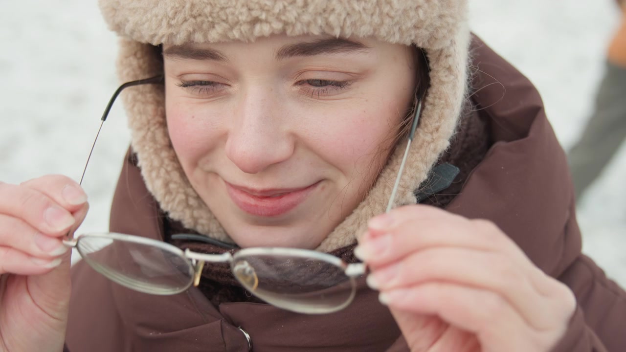 Close-up of young woman wearing shearling hat, round eyeglasses, high-collared brown padded coat and knit scarf as she lifts glasses with both hands, smiling gently in soft snowy daylight