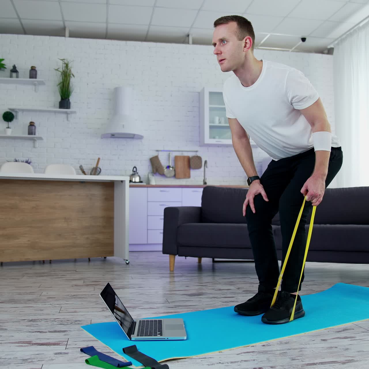 Concentrated man exercising with a rubber band at home. Young man doing working out while standing on a mat in the kitchen. Sportive lifestyle during quarantine