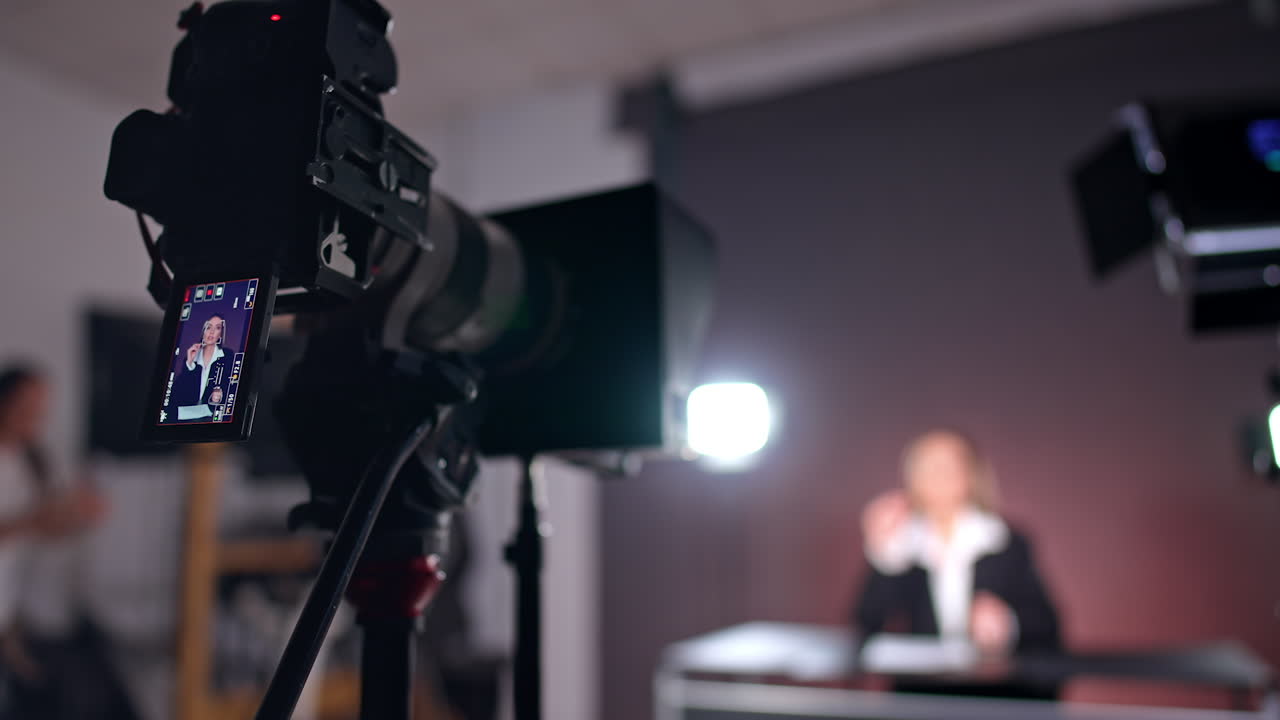 Camera on tripod is filming a lady sitting at backdrop. Close up. Low angle view. Video content creation in studio.