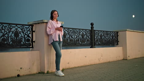 woman standing outdoors at dusk, leaning against decorative iron fence, wearing headphones and holding phone, soft evening lighting with moon visible in background