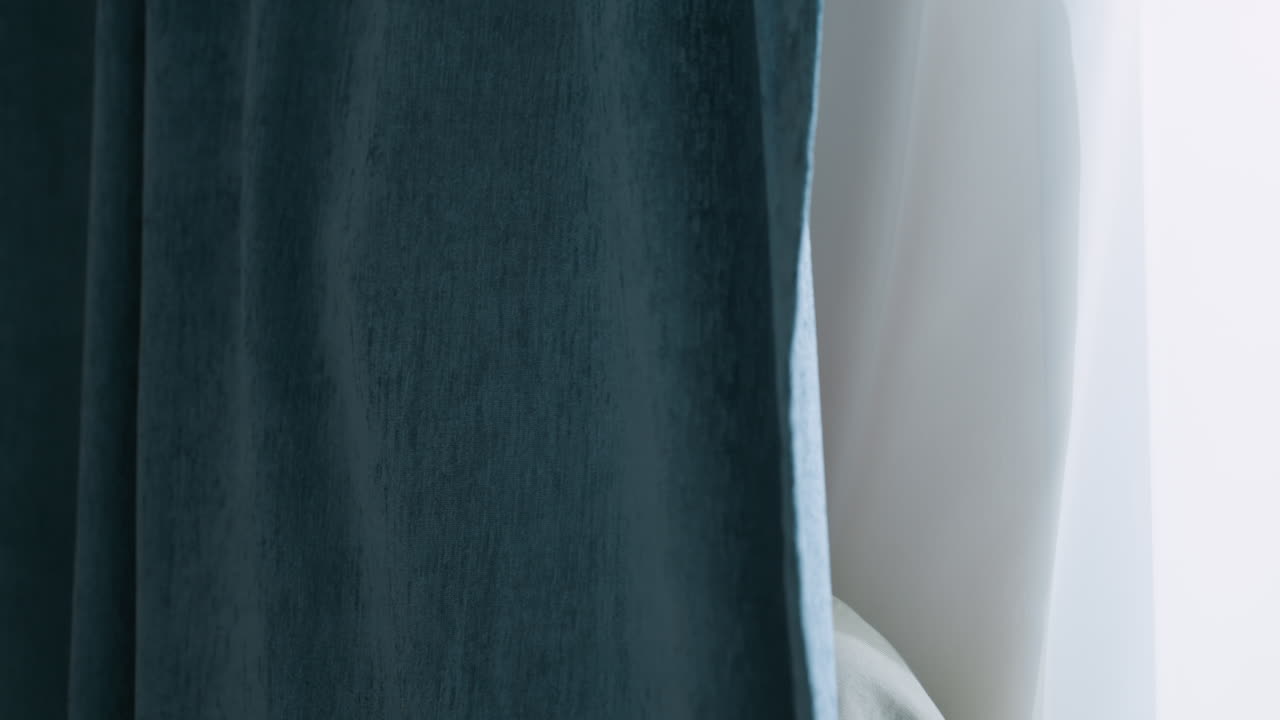 Teen boy stands partially hidden behind dark blue curtain near window with sheer white drapes, wearing calm expression in soft light, conveying thoughtful or reflective mood in quiet indoor scene