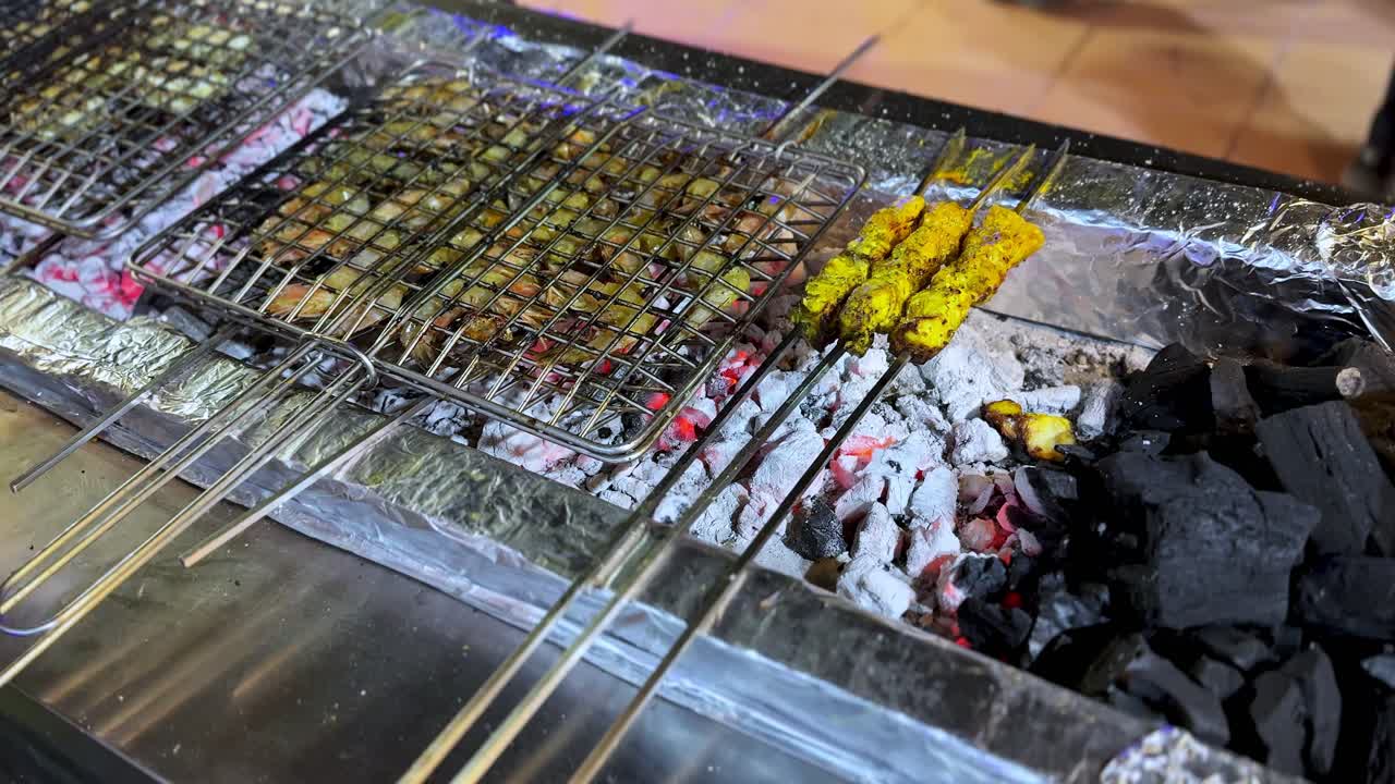 Close-up of a chef wearing black hygiene gloves grilling yellow marinated chicken skewers (Satay) on a wire rack over hot glowing charcoal embers and aluminum foil