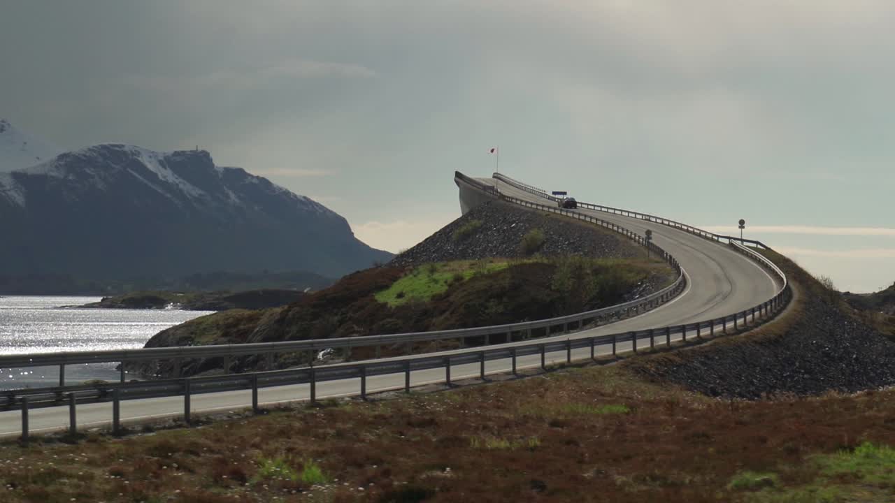 A view of the famous Atlantic Road in Norway. One of its'  curved bridges rises above the sea. It is overcast, with heavy clouds hanging above the water. A dark silhouette of a mountain on the horizon