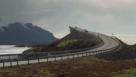 a view of the famous atlantic road in norway. one of its'  curved bridges rises above the sea. it is overcast, with heavy clouds hanging above the water. a dark silhouette of a mountain on the horizon
