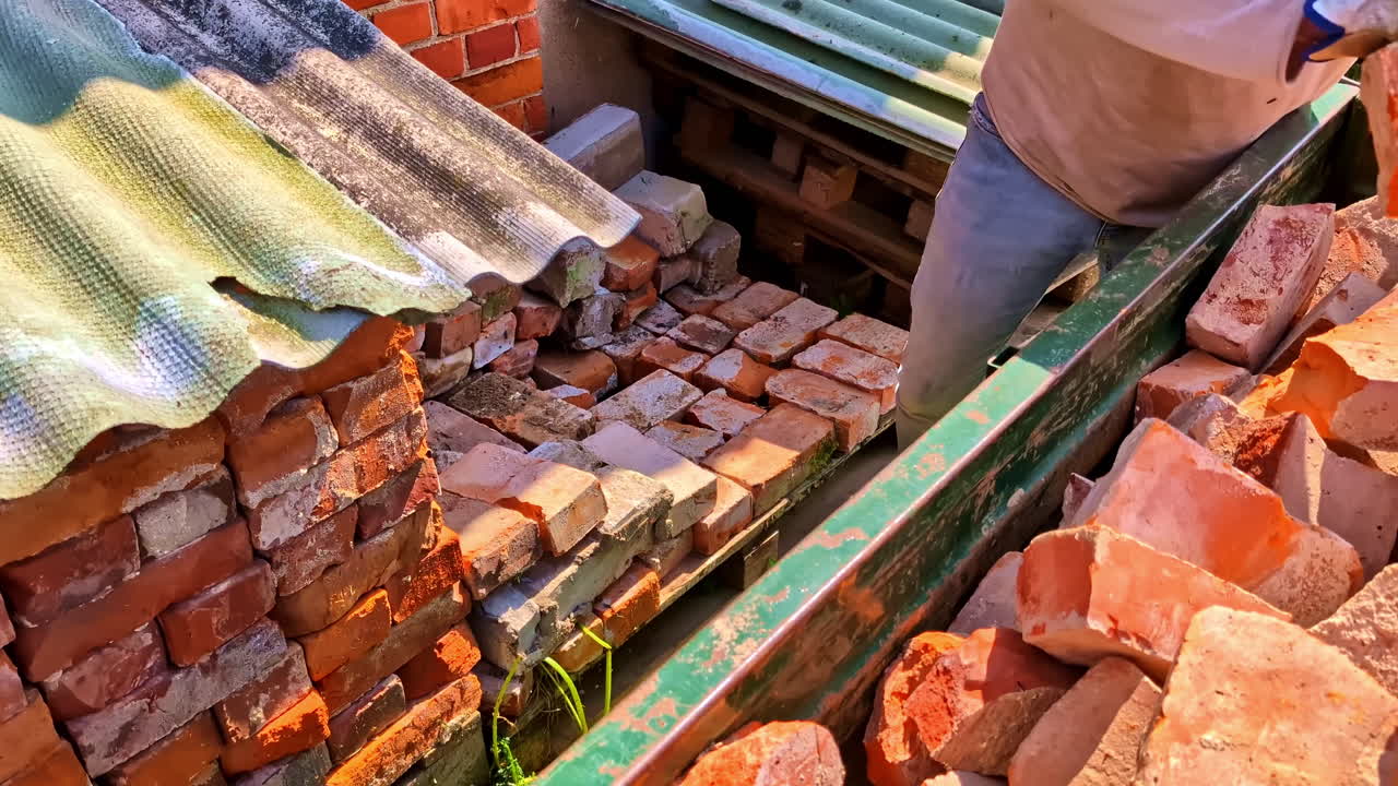 Man unloading bricks from trailer into stack covered with corrugated roof slate in sunlight