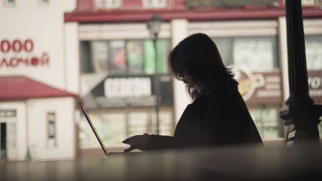 side view of woman typing on laptop outdoors with cascading hair, wearing a black coat, urban setting features soft-focus background of commercial buildings and streetlights