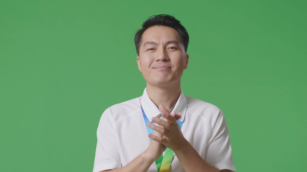 금메달을 달고 손을 치는 아시아 남자가 스튜디오의 녹색 스크린 배경에서 첫 번째 우승자로 자랑스럽게 승리하고 있습니다.