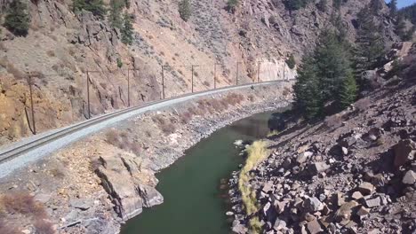 trucking-aerial-shot-of-a-railroad-track-following-an-emerald-green-river-with-power-lines-and-pine-trees-in-a-western-setting-with-lots-of-rocky-terrain-and-hills