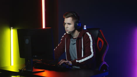 close up view of young serious man with headphones and playing a game on the computer in a room with colorful neon lamps on the wall 1